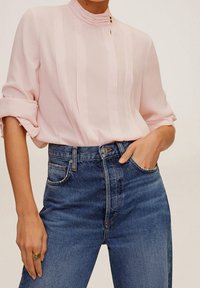 Helles rosa Bluse mit hohen Kragen und dezenten Streifen, kombiniert mit hoch taillierten blauen Jeans. Glatter Stoff, lässiger Schnitt.