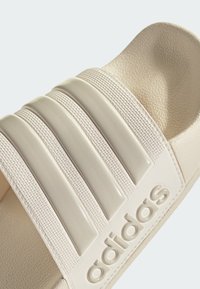 Sandalias de slide en color crema con tres tiras texturizadas y acanaladas, suave forro interior y logo en relieve. El material parece liso y duradero.