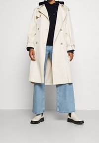 Gabardina beige con diseño de doble botonadura y grandes botones, llevada sobre un sudadera azul marino. Jeans de pierna ancha en azul claro y botas cortas crema.