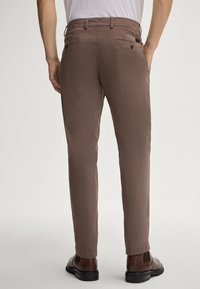 Pantaloni marroni, aderenti, con una texture liscia, due tasche posteriori con bottoni e un design a gamba affusolata. Indossati con scarpe eleganti scure.