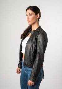 Schwarze Lederjacke mit einem kurvenartigen Design, Reißverschluss vorne, Druckknopfkragen und seitlichen Akzenten. Kombiniert mit hoch geschnittenen blauen Jeans.