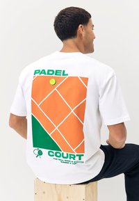 Maglietta bianca in cotone con una grafica sul retro comprendente elementi arancioni, verdi e bianchi, incluso il testo "PADEL COURT" e una pallina da tennis.