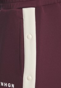 Bordo sporta bikses ar baltu sānu svītru un pogām, mīksta auduma tekstūra, un kabata kreisajā pusē. Akcenti ietver drukātus tekstus.