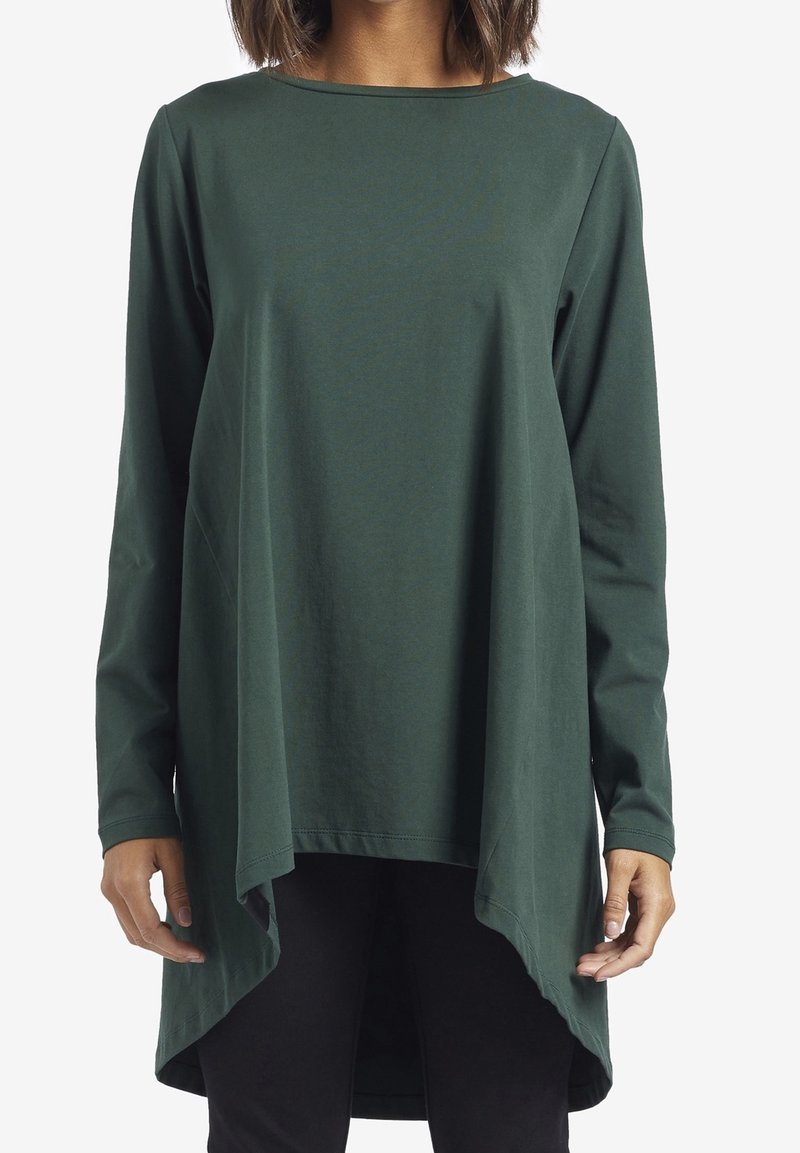 Maglia verde a maniche lunghe in tessuto morbido, con scollatura a barca, orlo asimmetrico e fit rilassato. Indossata sopra pantaloni neri.