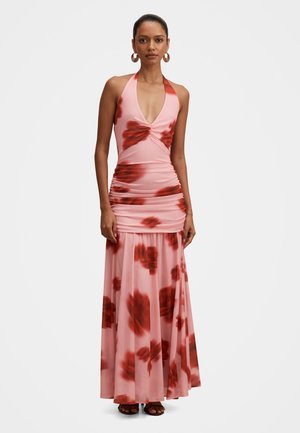 Naine, kes kannab varrukateta roosat halteri maxi kleiti punaste lillemustritega, kortsutatud vöökohaga ja suuri rõngaskõrvarõngaid, seisab ühevärvilise tausta ees.