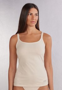 Débardeur beige clair en tissu doux avec des fines bretelles, un col carré et une silhouette ajustée. Étiquette de logo subtile en bas de l'ourlet.