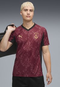Maronen Fußballtrikot mit schwarzen kurzenÄrmeln; verfügt über ein kreisförmiges Muster und ein goldenes Logo; V-Ausschnitt-Design; leichtes, glattes Stoffmaterial.