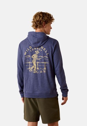 Man wearing blue hoodie with hood up and beige cowboy skeleton graphic and "Wild Hell Cowboy" text on the back, paired with olive shorts.