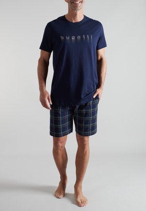 Mann trägt ein marineblaues T-Shirt mit dem Schriftzug "bugatti" und dunkelblaue karierte Shorts, steht barfuß vor schlichtem Hintergrund.