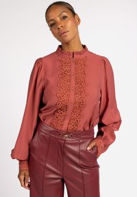 Rostfarbendes Blusenshirt mit hohem Rüschkragen, der auf der Vorderseite über feine Spitzenverzierungen verfügt, sowie mit Ballonärmeln.