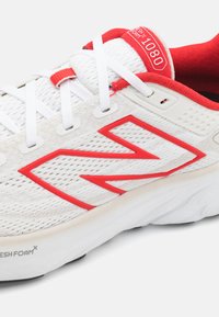 Sapatilha desportiva branca com pormenores em vermelho, parte superior em malha respirável, sola espessa, logótipo "N" em destaque e colarinho acolchoado para conforto.