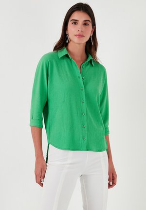Green button-up shirt with a textured finish, roll-up sleeves, a collared neck, and a slightly curved hem. Worn with white pants.