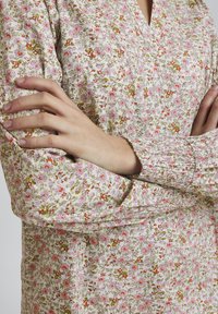 Blomstret kjole med en lys beige baggrund, prydet med pink og grønne blomster. Løs pasform, v-hals og smockede ærmer. Teksturen ser blød ud.