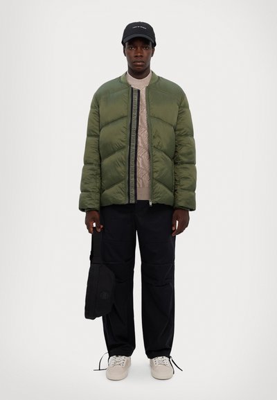 Veste en duvet verte avec coutures en chevron, pull en tricot beige, pantalon noir et baskets claires ; portant un sac noir.