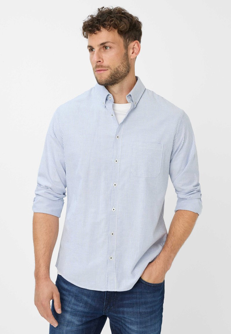 Man met krullend haar die een lichtblauw gestreept overhemd over een wit T-shirt draagt, blauwe jeans, rechterhand in de zak, kijkend naar links.