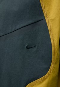 Nike sportinis marškinėlis su tamsiai žaliu ir garstyčių geltonu raštu, lygia tekstūra, plokščiais siūlais ir išsiuvinėtu juodu logotipu.