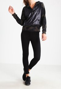 Mujer vestida con una sudadera de camuflaje, un chaleco negro brillante, leggings negros y zapatillas negras, de pie con una pierna ligeramente doblada.