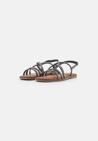 Sandali marroni con cinturini e accenti metallici, suola piatta e cinturino alla caviglia regolabile, caratterizzati da un design con punta aperta e parte superiore testurizzata.