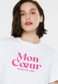 Weißes T-Shirt mit kurzen Ärmeln, das vorne mit fettem pinkem Text "Mon Cœur SENSES.THE LABEL" versehen ist. Glatte Baumwollqualität.