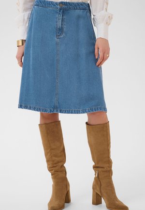 Vrouw die een blauwe denim rok tot op de knie draagt, een witte blouse en beige suède kniehoge laarzen met hakken en zijritsen.