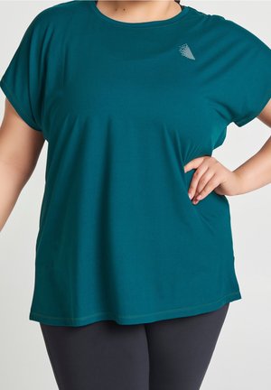 Femme portant un haut ample à manches courtes bleu sarcelle et un pantalon foncé, debout avec une main sur la hanche devant un fond uni.