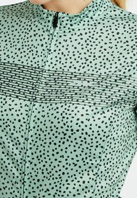 Veste vert menthe en tissu texturé avec un motif de taches noires et des rayures horizontales noires sur la poitrine. Fermeture par zip au cou.