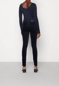 Långärmad marinblå topp med vid halsringning, kombinerad med åtsittande mörka jeans. Modellen bär bruna klacksandaler och visar de eleganta bakdetaljerna på jeansen.