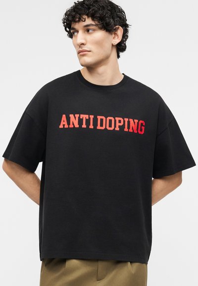 Νεαρός άνδρας με σγουρά μαλλιά που φοράει μαύρο φαρδύ T-shirt με κόκκινο κείμενο «ANTI DOPING», στέκεται με τα χέρια πίσω από την πλάτη του.