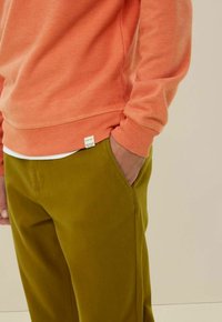 Koraalkleurige sweatshirt met ribgebreide manchetten, gecombineerd met olijfgroene broeken met een zijzak. De stof lijkt zacht met een lichte textuur.