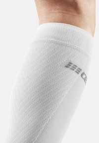 Weiße knielange Kompressionssocke mit strukturiertem Strickmuster und dezentem Branding an der Seite. Verfügt über eine glatte Stoffoberfläche.