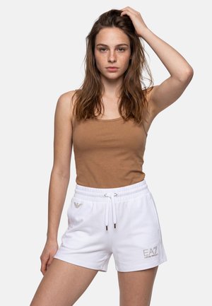 Giovane donna con lunghi capelli castani, canottiera marrone e pantaloncini bianchi EA7 Emporio Armani, che posa con una mano sulla testa.
