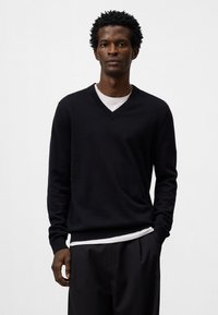 Maglione nero a scollo a V realizzato in tessuto morbido, con maniche lunghe e vestibilità rilassata, indossato sopra una t-shirt bianca a girocollo.