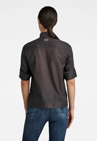 Femme aux cheveux bruns attachés en une queue de cheval basse, portant un t-shirt noir à manches courtes retroussées et un jean en denim bleu, vue de dos.