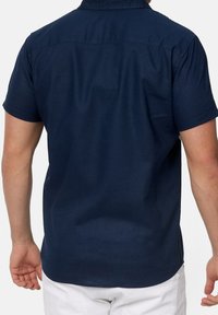 Camisa de manga corta azul marino con cuello abotonado, textura suave, dobladillo recto y una única costura en el yugo trasero. Diseño simple y minimalista.