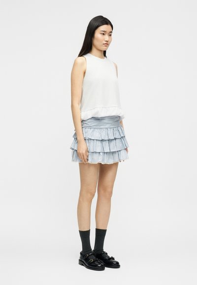 Jeune femme debout portant un haut blanc sans manches, une mini-jupe à volants superposés bleu clair, des chaussettes noires et des mocassins noirs sur un fond uni.