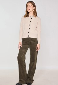 Cardigan beige en tricot avec des boutons noirs, col rond et texture côtelée, associé à un pantalon en velours côtelé évasé vert olive. Chaussures noires.