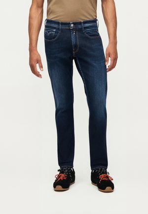 Mand iført mørkeblå slim-fit jeans og sorte sneakers med røde og hvide snørebånd, stående mod en hvid baggrund.