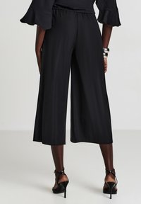 Schwarze weit geschnittene Culottes mit einem elastischen Bund, aus einem glatten Stoff gefertigt. Die Säume sind gerade geschnitten und werden mit schwarzen Riemchen-High Heels kombiniert.