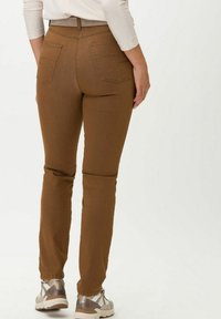Hnědé slim-fit kalhoty s hladkým povrchem, které mají dvě zadní kapsy a střední pas. V kombinaci se světlou obuví.