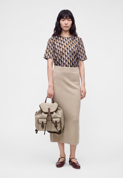 Femme portant un haut à motif géométrique, une jupe midi beige, des ballerines bordeaux, tenant un sac à dos beige par sa poignée sur un fond uni.