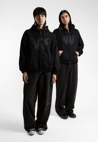 Le felpe nere con cappuccio e zip presentano una stampa grafica sul petto. Pantaloni neri dal taglio comodo e calzature di contrasto completano il look.