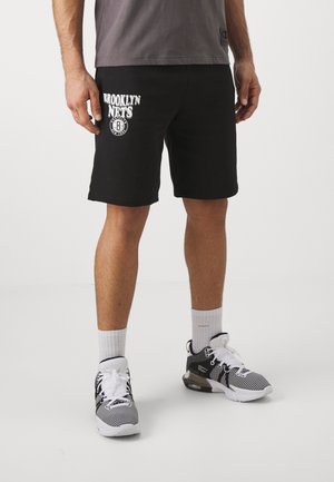 Mężczyzna w czarnych krótkich spodenkach Brooklyn Nets, białych skarpetkach i czarno-białych butach sportowych, stojący na jednolitym tle.