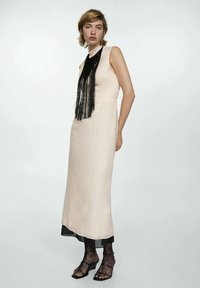Cremefarbenes ärmelloses Kleid mit hohem Kragen, verziert mit einer schwarzen Fransen-Halskette. Schwarze Spitzenstrumpfhosen und Absatzsandalen runden den Look ab.