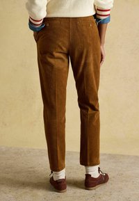 Brūnas kordurojas bikses ar taisnu staru griezumu, divām aizmugures kabatām un mīkstu tekstūru. Kombinētas ar brūnām kurpēm un baltām zeķēm.