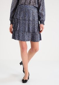 Falda azul de volantes con un sutil patrón de puntos metálicos, textura plisada y cinturilla elástica; combinada con zapatos de charol negros.