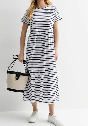Kvinde i hvid og sort stribet midi-kjole, der holder en beige og sort håndtaske, iført hvide sneakers, stående mod en hvid baggrund.