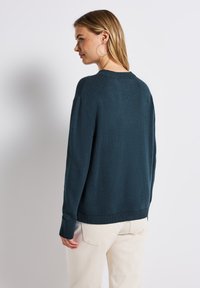 Türkisfarbener Strickpullover mit Rundhalsausschnitt, überschnittenen Schultern und gerippten Bündchen. Stilisiert mit hellen Hosen.
