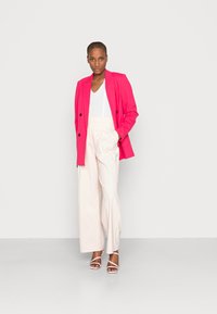 Blazer rosa duplo sobre uma blusa branca, combinado com calças largas em tom creme e sandálias rosa pálido, contra um fundo neutro.