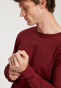 Burgundowa koszula z długim rękawem, wykonana z miękkiego materiału, z okrągłym dekoltem, dopasowanymi mankietami i subtelnym detalem z logo na piersi.