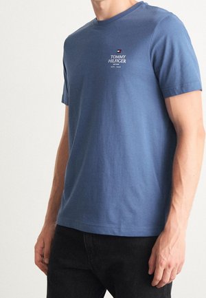 Mężczyzna w niebieskiej koszulce Tommy Hilfiger z logo i napisem na lewej piersi, w czarnych spodniach, stojący na białym tle.